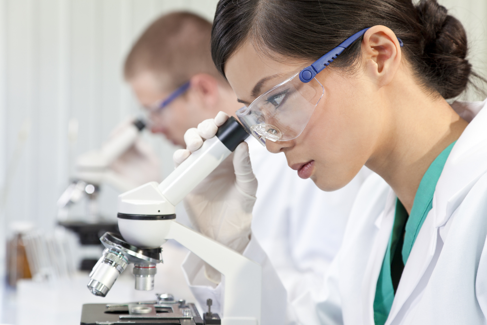 A Chinese Asian female medical or scientific researcher or doctor using a microscope in a laboratory with her colleague out of focus behind her.
